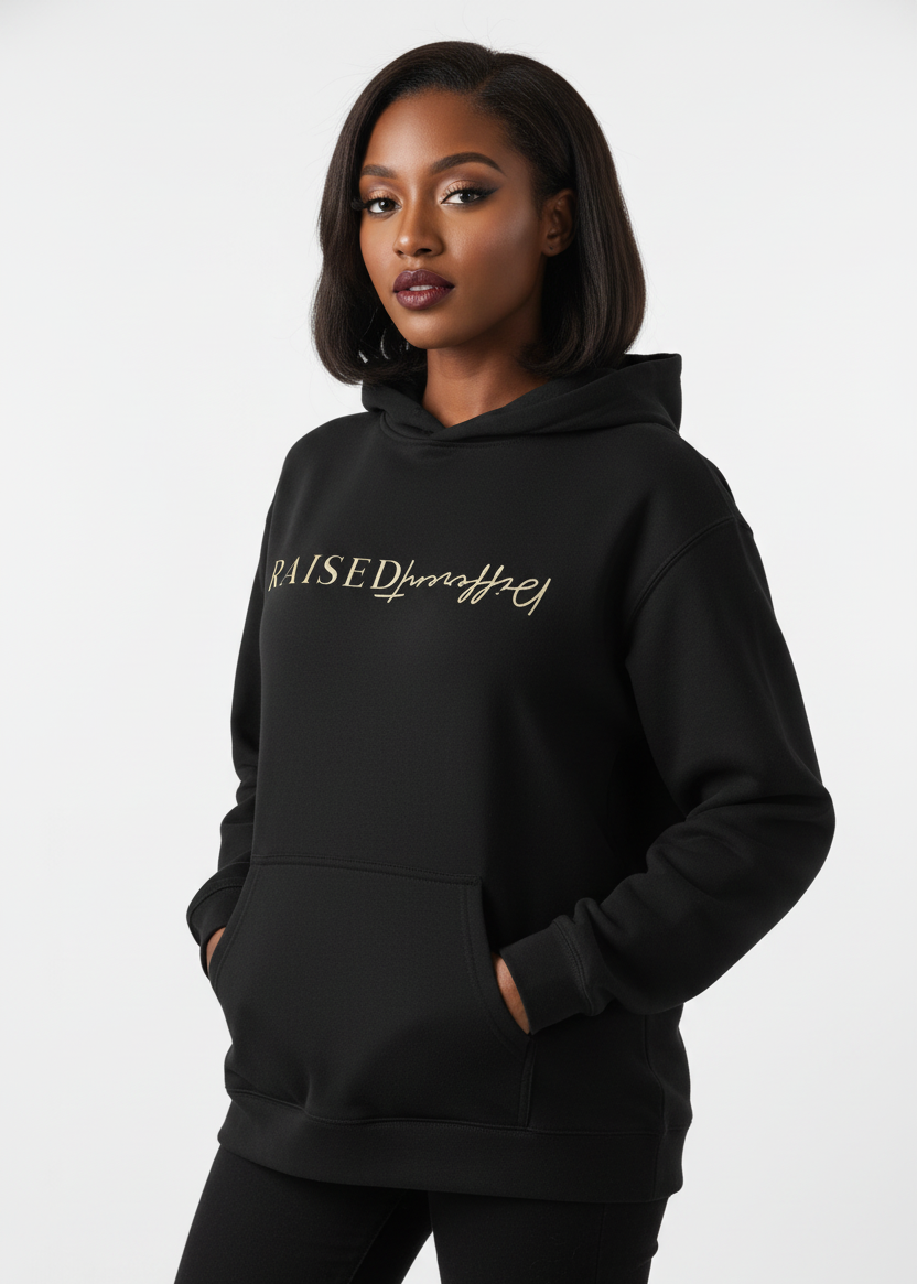 Woman wearing a black affirmation hoodie with 'Raised Different' text, standing against an urban background.