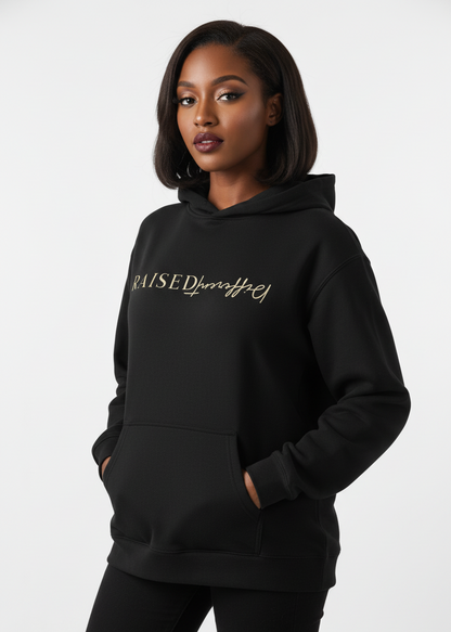 Woman wearing a black affirmation hoodie with 'Raised Different' text, standing against an urban background.
