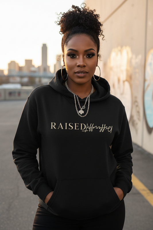 Woman wearing a black affirmation hoodie with 'Raised Different' text, standing against an urban background.