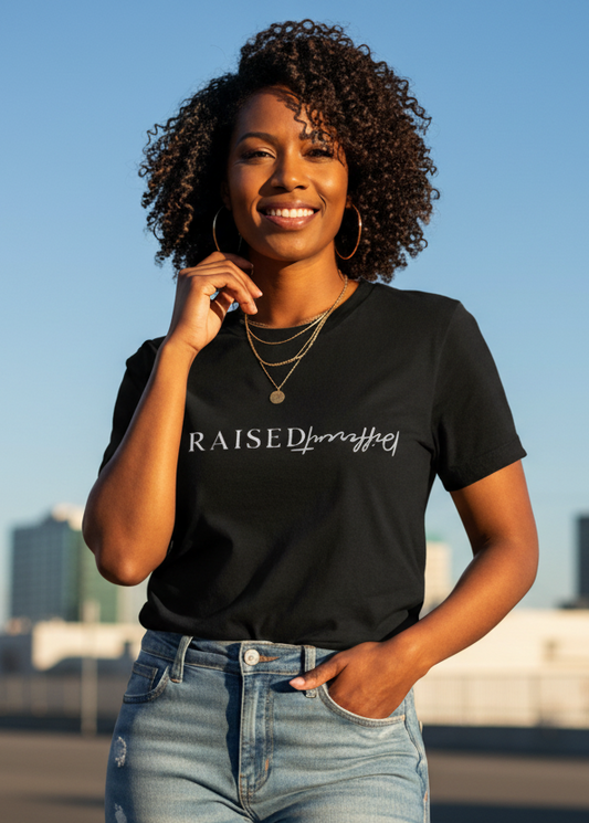 Brown woman wearing a black t-shirt with 'Raised Different' text, standing outdoors with a cityscape in the background.