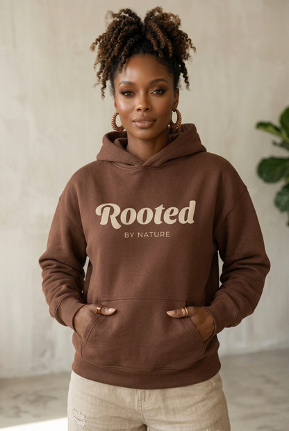 A woman wearing a brown pullover hoodie with 'Rooted BY NATURE' printed in beige across the chest, standing indoors against a light textured wall.