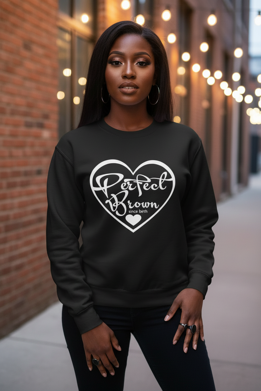 A woman wearing a black crewneck sweatshirt with a white heart graphic that reads 'Perfect Brown Love All' standing on a city sidewalk next to a brick wall with warm string lights in the background.