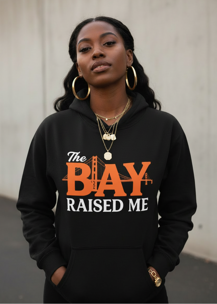 A woman wearing a black pullover hoodie with a graphic print that reads 'The BAY RAISED ME' in orange and white, standing against a concrete wall.
