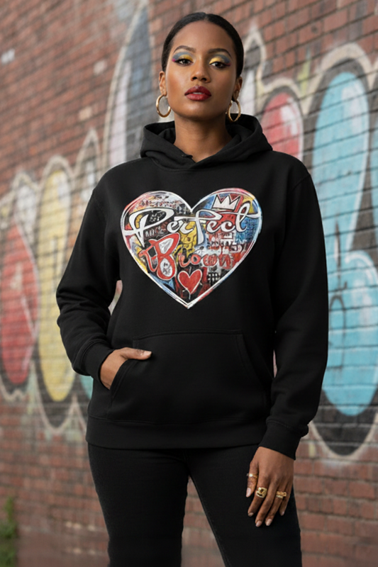 A woman wearing a black hoodie with a large, colorful graffiti-style heart graphic on the chest, standing in front of a brick wall with graffiti.