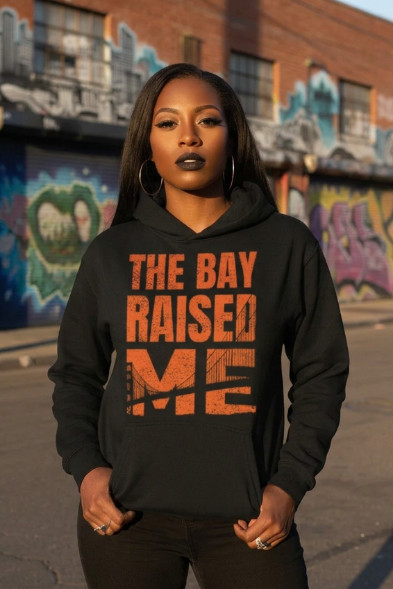 A woman wearing a black hoodie with the phrase 'THE BAY RAISED ME' printed in distressed orange lettering, standing in front of a graffiti-covered brick wall.