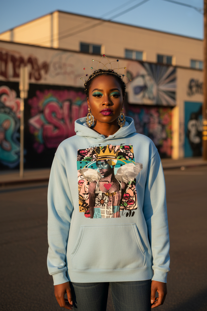 A woman stands outdoors in front of a graffiti-covered wall, wearing a light blue pullover hoodie with a large colorful graphic print on the front. The print shows a stylized portrait with pink, gold, and white elements.