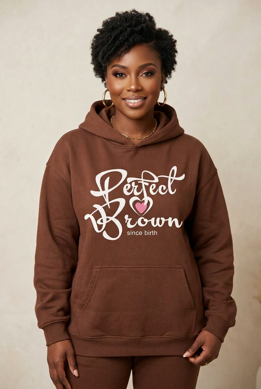 A woman wearing a brown pullover hoodie with a front kangaroo pocket and a graphic that reads 'Perfect Brown since birth' in white script with a small pink heart, posed against a neutral beige background.