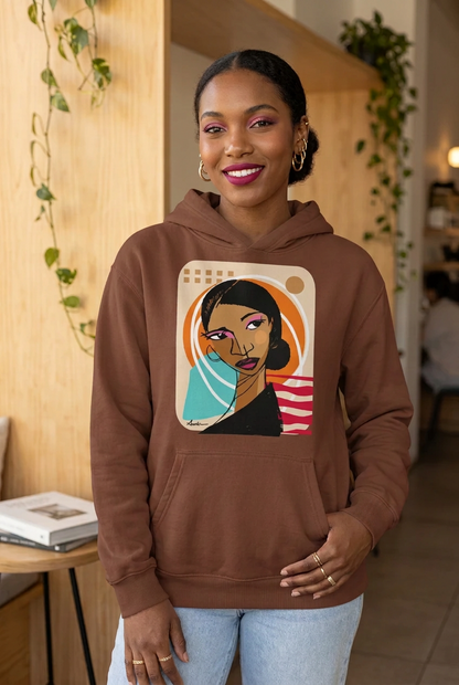 A woman wearing a brown pullover hoodie with a colorful graphic print of a woman's face on the chest, standing in a warmly lit indoor space with wooden walls and hanging greenery.