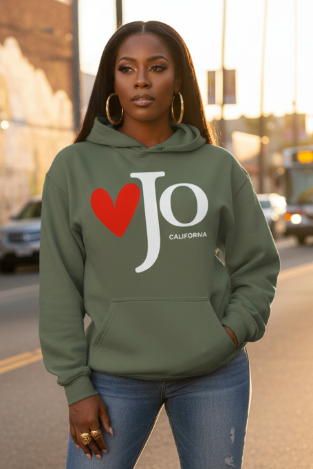 A woman wearing an olive green hoodie with a red heart and white 'Jo California' graphic, standing on a city street at sunset.
