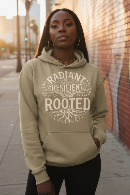 A woman wearing a beige hoodie with a white graphic print that reads 'RADIANT RESILIENT ROOTED' with a tree design, standing on a city sidewalk next to a brick wall.