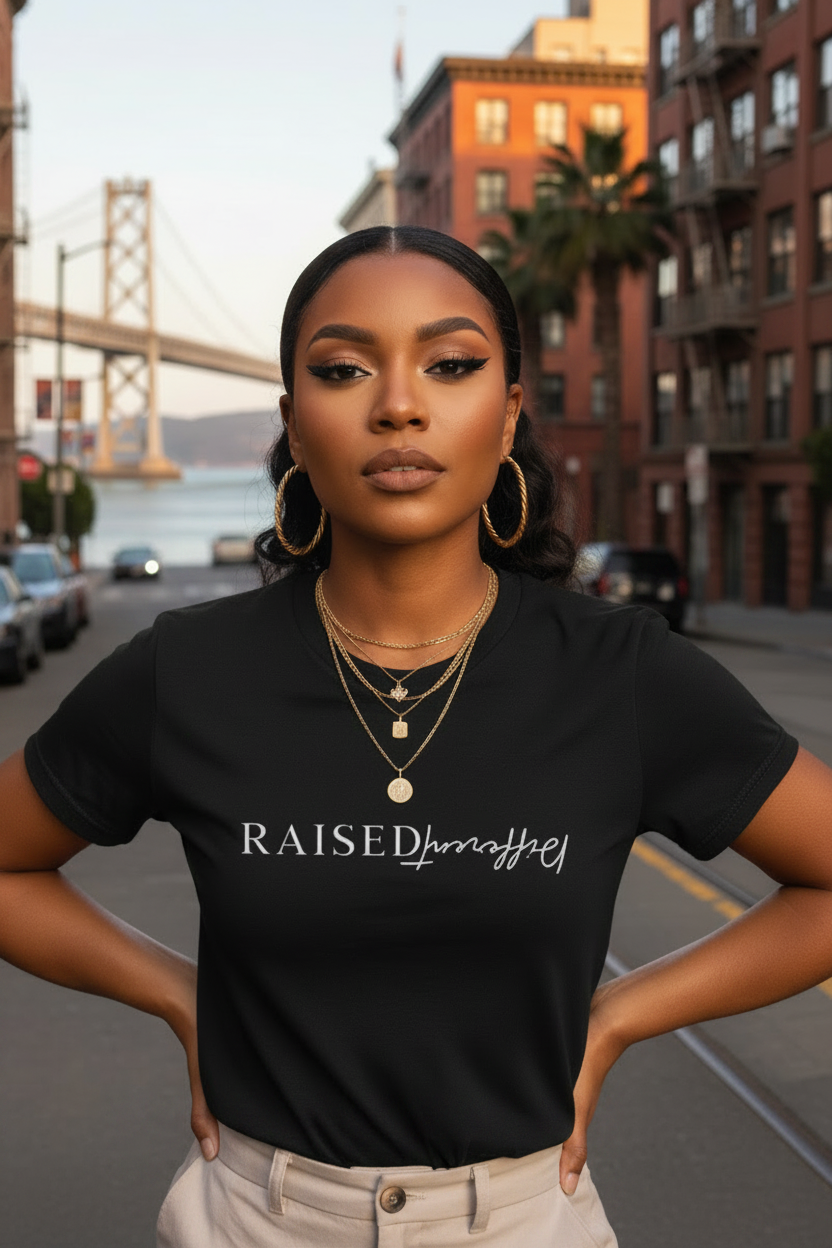 A woman stands on a city street wearing a black short-sleeve T-shirt with the text 'RAISED by Affair' printed across the chest, paired with light beige pants and layered gold necklaces. The background shows a blurred urban scene with a bridge and buildings.