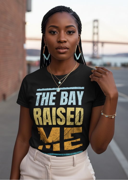 A woman wearing a black graphic t-shirt with the text 'THE BAY RAISED ME' in distressed light blue and yellow lettering, paired with cream pants, standing outdoors with a bridge and brick wall in the background.