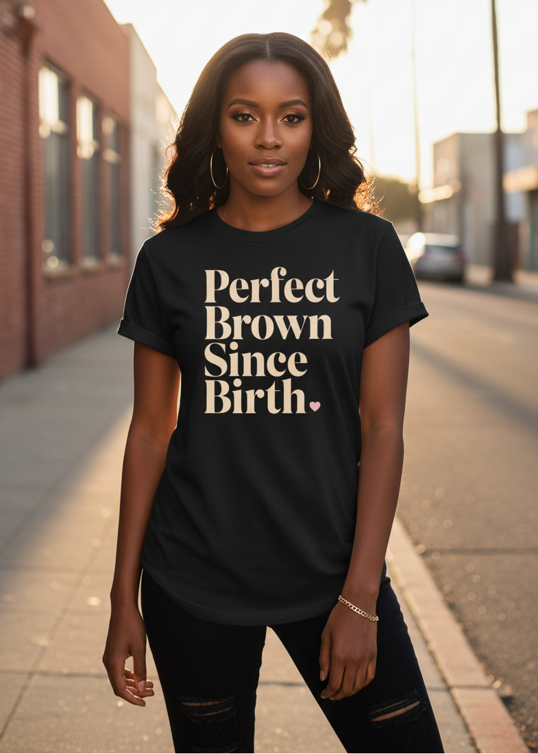 A woman stands on a city sidewalk wearing a black graphic T-shirt with the phrase 'Perfect Brown Since Birth.' printed in beige serif text across the front, paired with black ripped jeans.