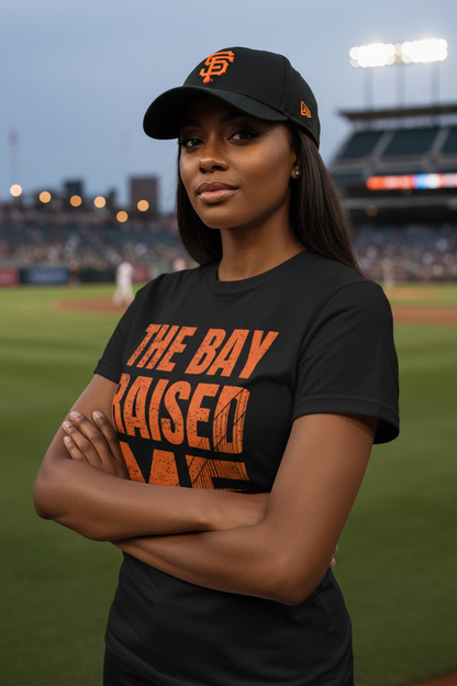 A woman stands on a baseball field wearing a black San Francisco Giants cap and a black T-shirt with orange text reading 'THE BAY RAISED ME'. The background shows a blurred stadium at dusk.