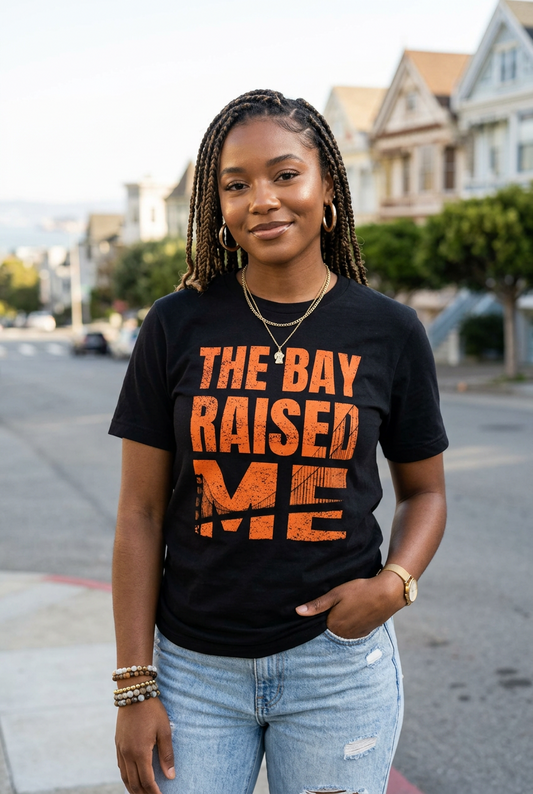A woman stands on a city street wearing a black graphic T-shirt with distressed orange text reading 'THE BAY RAISED ME' and light blue jeans. The background shows blurred houses and trees.