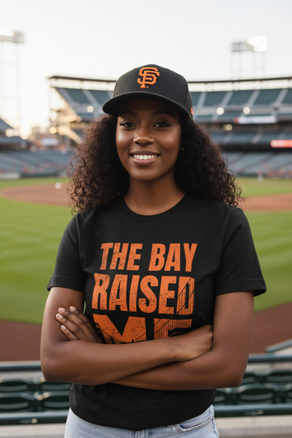 A smiling woman stands on a baseball field wearing a black San Francisco Giants cap and a black T-shirt with distressed orange text reading 'THE BAY RAISED ME'. The stadium is blurred in the background.