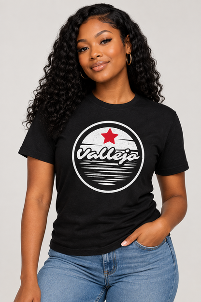 A woman with long, curly black hair wearing a black crewneck t-shirt with a circular 'Vallejo' graphic on the chest, paired with blue jeans, against a solid light gray background.