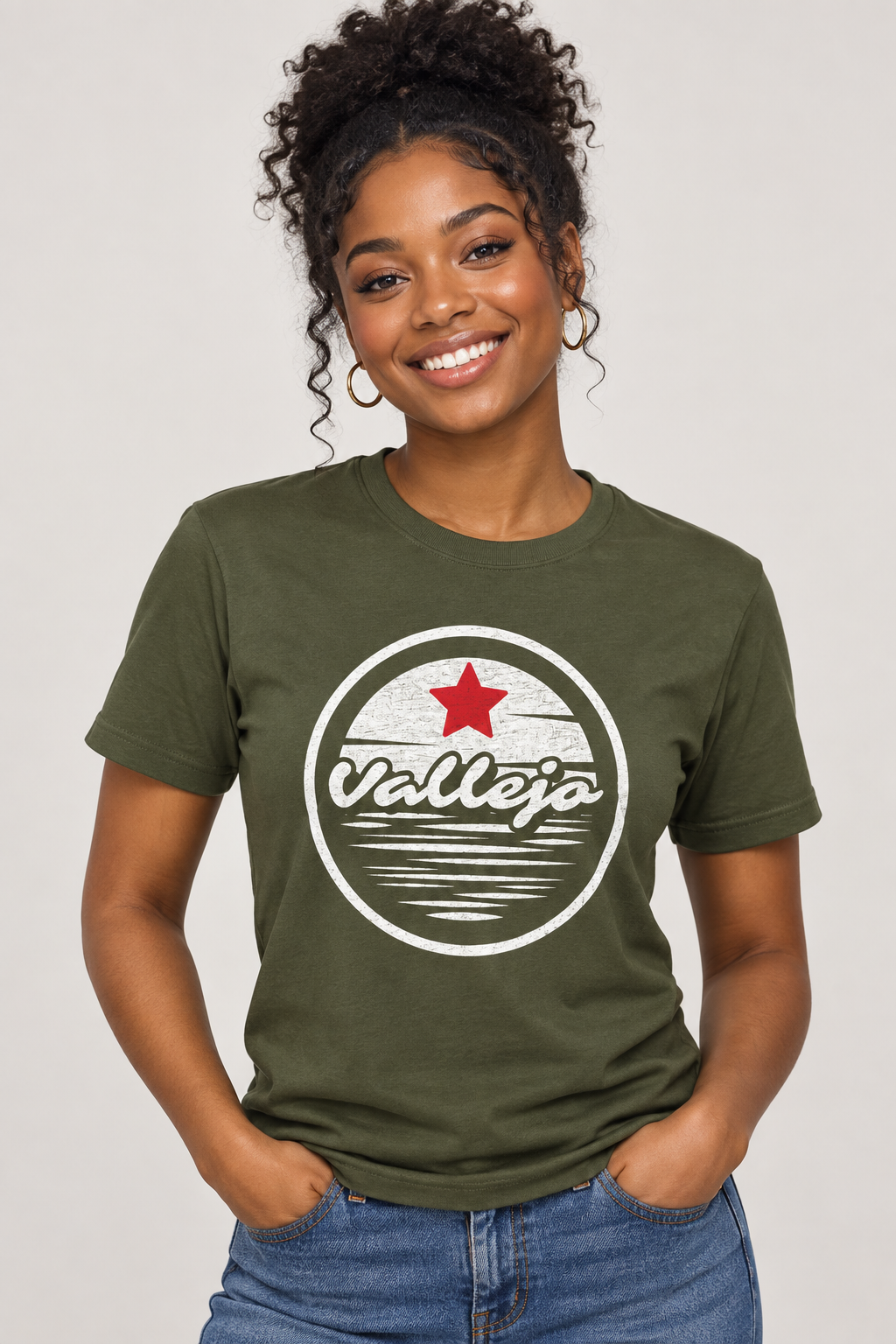 A woman wearing an olive green t-shirt with a circular 'Vallejo' graphic on the chest, paired with blue jeans, against a plain light gray background.