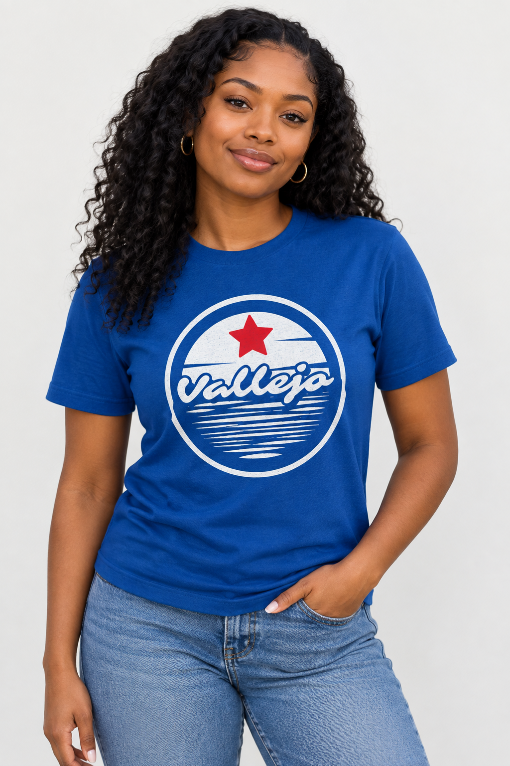 A woman with curly black hair wearing a royal blue crewneck t-shirt with a circular 'Valleja' graphic on the chest, paired with light-wash blue jeans, against a light gray studio background.