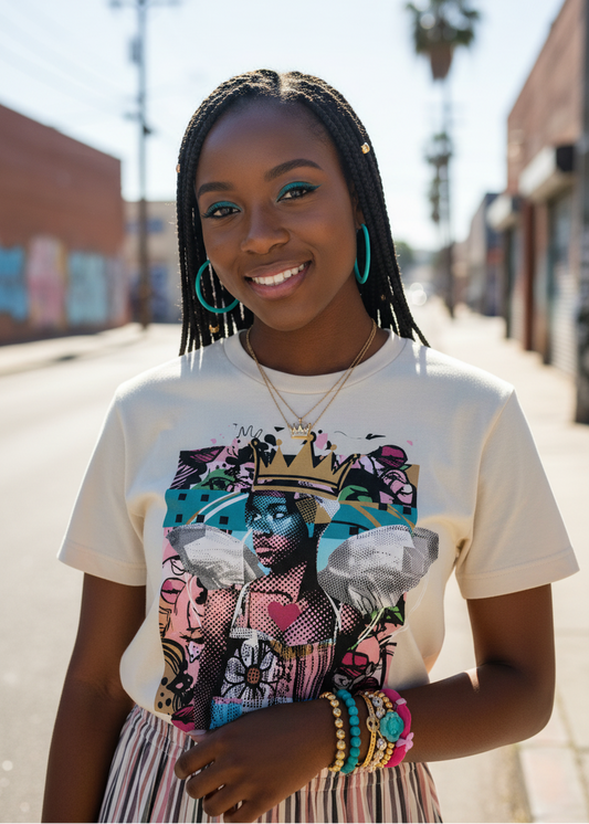 A woman stands on a sunny city street wearing a cream graphic T-shirt with a colorful illustrated print, striped pants, and turquoise hoop earrings.